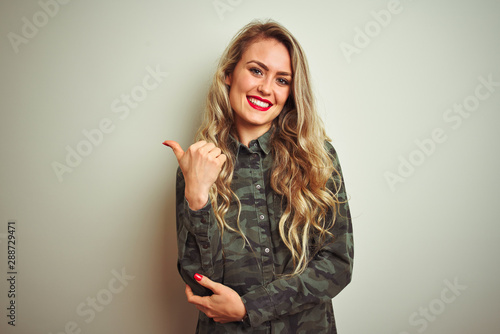 Young beautiful woman wearing military camouflage shirt over white isolated background smiling with happy face looking and pointing to the side with thumb up.