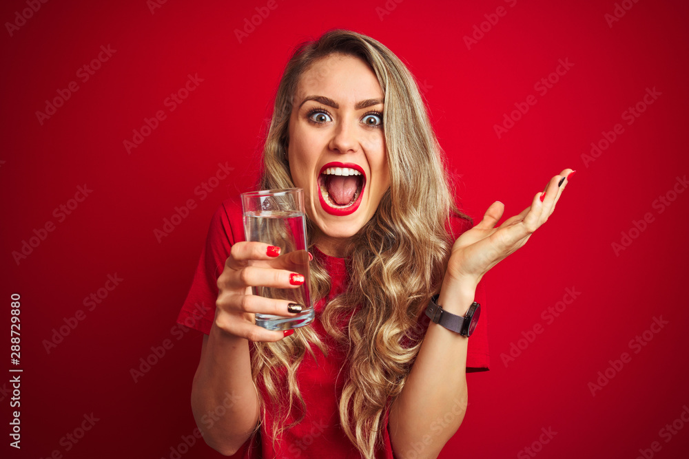 Young beautiful woman drinking a glass of water over red isolated background very happy and excited, winner expression celebrating victory screaming with big smile and raised hands Young beautiful woman drinking a glass of water over red isolated background very happy and excited, winner expression celebrating victory screaming with big smile and raised hands