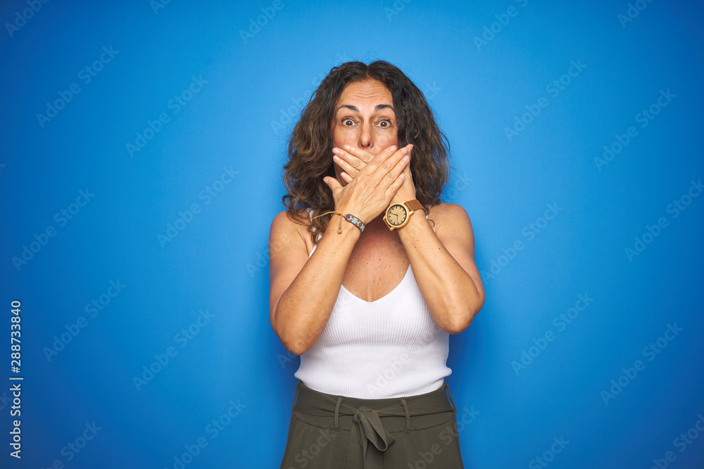 Middle age senior woman with curly hair standing over blue isolated background shocked covering mouth with hands for mistake. Secret concept. Middle age senior woman with curly hair standing over blue isolated background shocked covering mouth with hands for mistake. Secret concept.