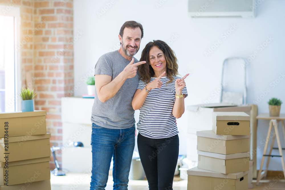 Middle age senior couple moving to a new home with boxes around smiling and looking at the camera pointing with two hands and fingers to the side. Middle age senior couple moving to a new home with boxes around smiling and looking at the camera pointing with two hands and fingers to the side.