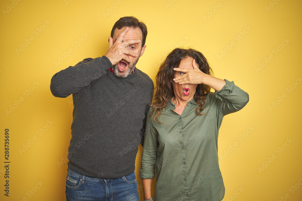 Beautiful middle age couple over isolated yellow background peeking in shock covering face and eyes with hand, looking through fingers with embarrassed expression. Beautiful middle age couple over isolated yellow background peeking in shock covering face and eyes with hand, looking through fingers with embarrassed expression.