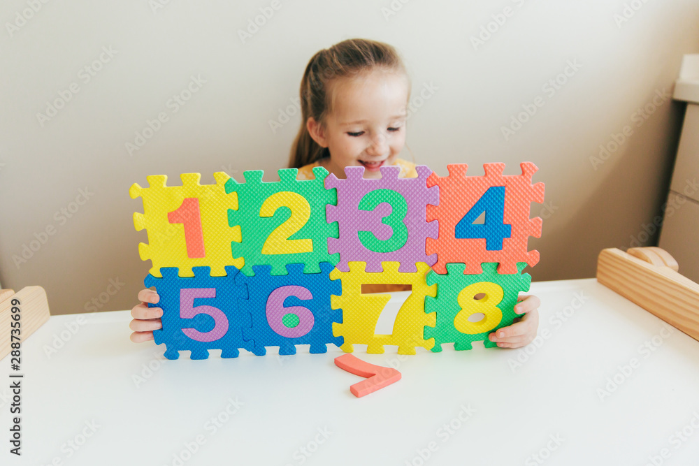 Fun, joy and happy childhood and education concept. White little girl in casual yellow dress holds assembled bright, multi-colored puzzle with numbers from one to eight Fun, joy and happy childhood and education concept. White little girl in casual yellow dress holds assembled bright, multi-colored puzzle with numbers from one to eight
