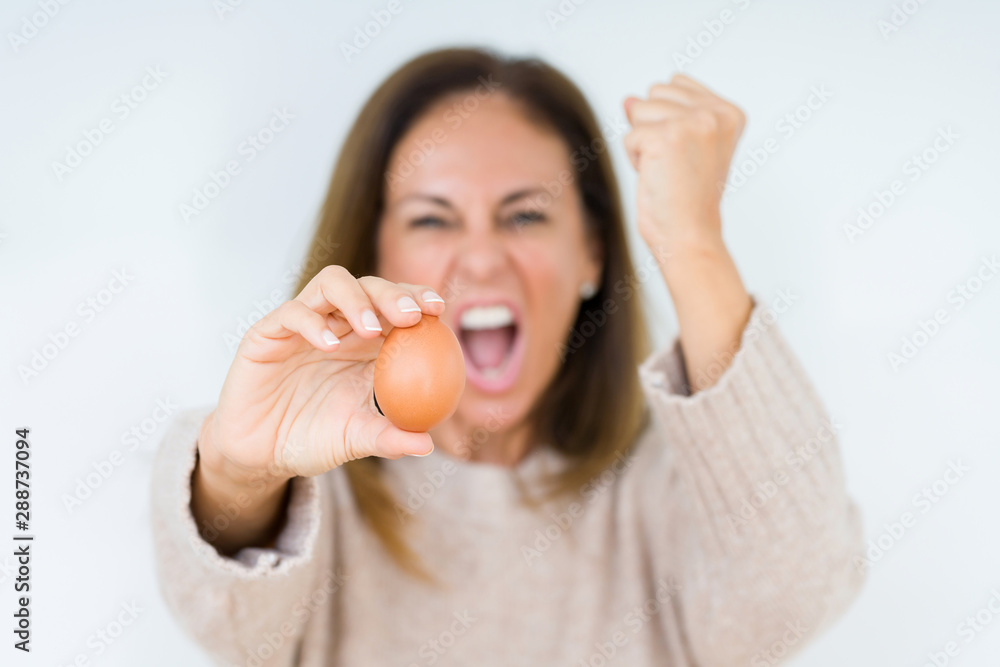 Middle age woman holding fresh egg over isolated background annoyed and frustrated shouting with anger, crazy and yelling with raised hand, anger concept Middle age woman holding fresh egg over isolated background annoyed and frustrated shouting with anger, crazy and yelling with raised hand, anger concept
