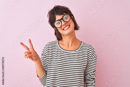 Young beautiful woman wearing striped t-shirt and glasses over isolated pink background smiling with happy face winking at the camera doing victory sign. Number two.