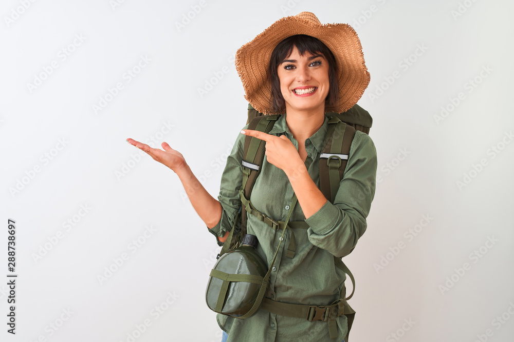 Hiker woman wearing backpack hat and water canteen over isolated white background amazed and smiling to the camera while presenting with hand and pointing with finger. Hiker woman wearing backpack hat and water canteen over isolated white background amazed and smiling to the camera while presenting with hand and pointing with finger.