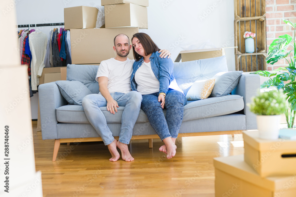 Young couple sitting on the sofa arround cardboard boxes moving to a new house Relaxed with serious expression on face. Simple and natural looking at the camera. Young couple sitting on the sofa arround cardboard boxes moving to a new house Relaxed with serious expression on face. Simple and natural looking at the camera.