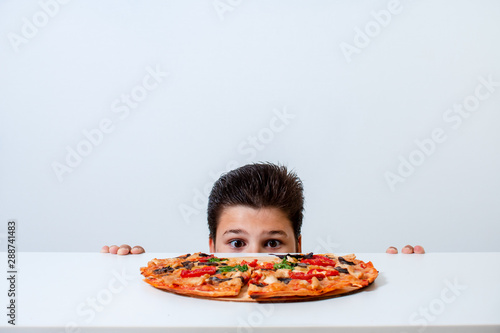 The boy eats Italian pizza. Looks out from behind the table
