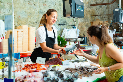 Polite  female worker of fish shop in apron offering fresh raw salmon to woman customer