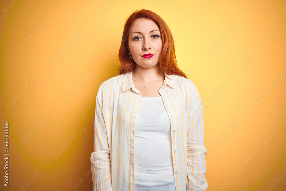 Obraz premium Beautiful redhead woman wearing striped shirt standing over isolated yellow background with serious expression on face. Simple and natural looking at the camera.