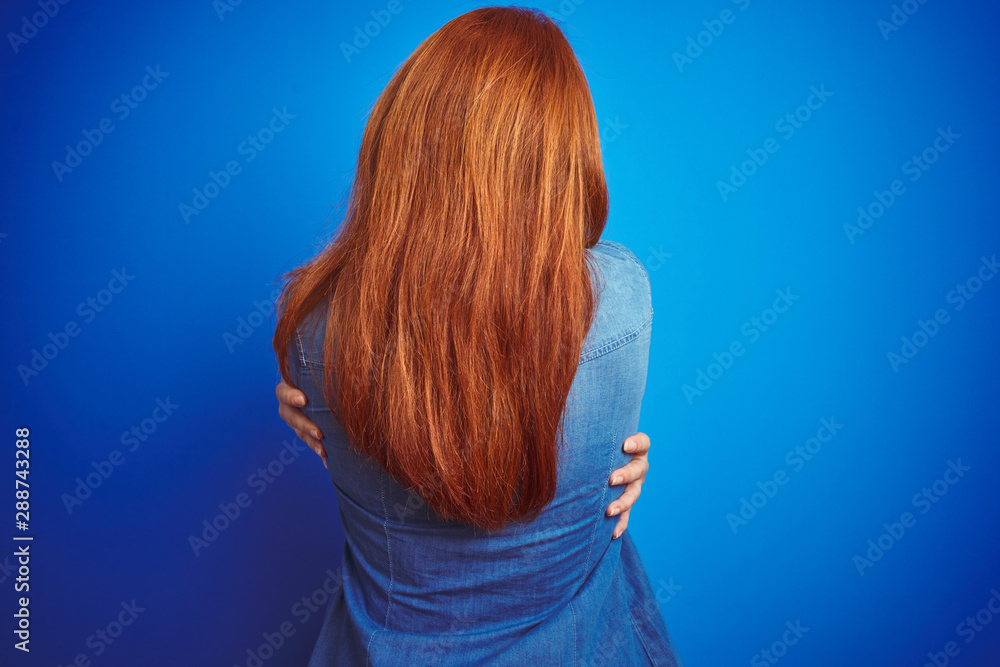 Young beautiful redhead woman wearing denim shirt standing over blue isolated background Hugging oneself happy and positive from backwards. Self love and self care - obrazy, fototapety, plakaty 