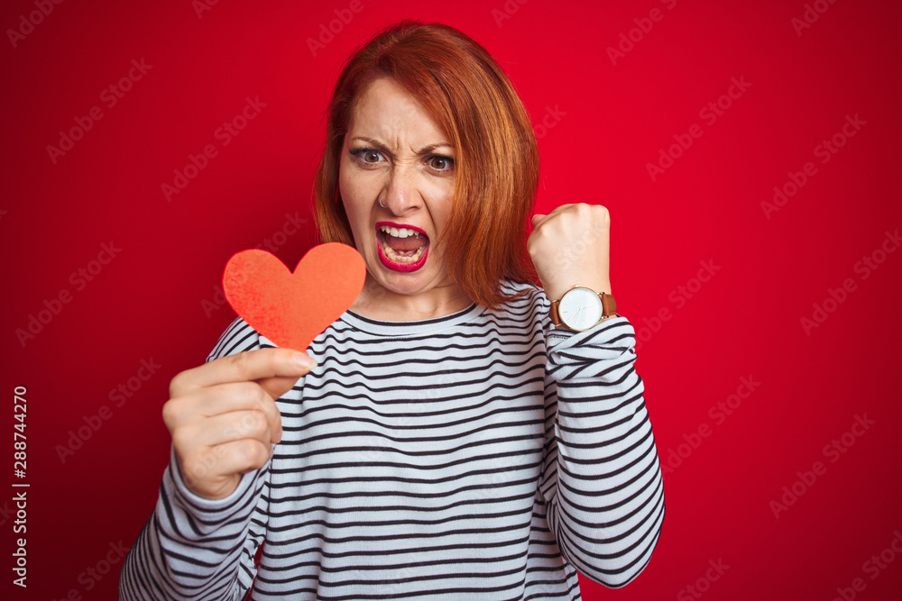 Beautiful redhead romantic woman holding heart standing over isolated red background annoyed and frustrated shouting with anger, crazy and yelling with raised hand, anger concept Beautiful redhead romantic woman holding heart standing over isolated red background annoyed and frustrated shouting with anger, crazy and yelling with raised hand, anger concept