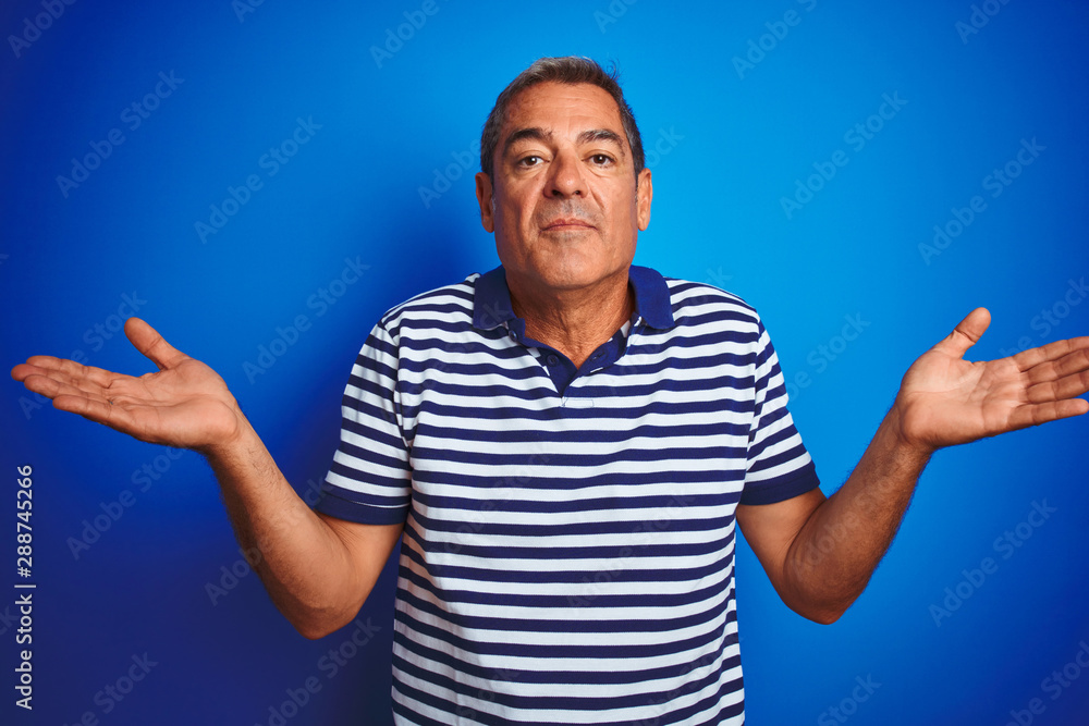 Handsome middle age man wearing striped polo standing over isolated blue background clueless and confused expression with arms and hands raised. Doubt concept. Handsome middle age man wearing striped polo standing over isolated blue background clueless and confused expression with arms and hands raised. Doubt concept.