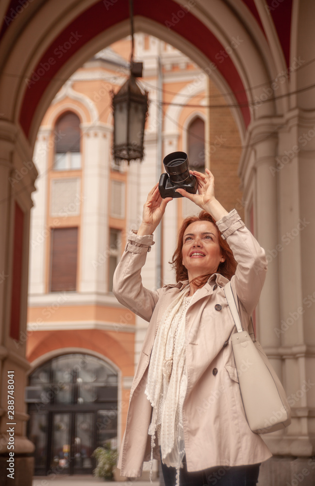 One mature woman, front view, holding a camera, photographing landmark in a city, old European architecture.
