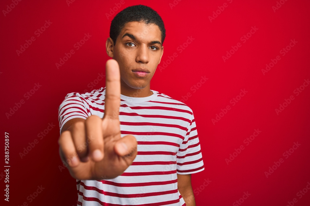 Young handsome arab man wearing striped t-shirt over isolated red background Pointing with finger up and angry expression, showing no gesture