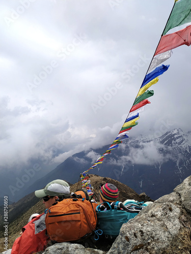 Tourists rests on the top of a mountain in the Himalayas, prayer flags develops over their heads. Nature, religion, healthy lifestyle, outdoors, east asian culture, hiking, travel and tourism concept.
