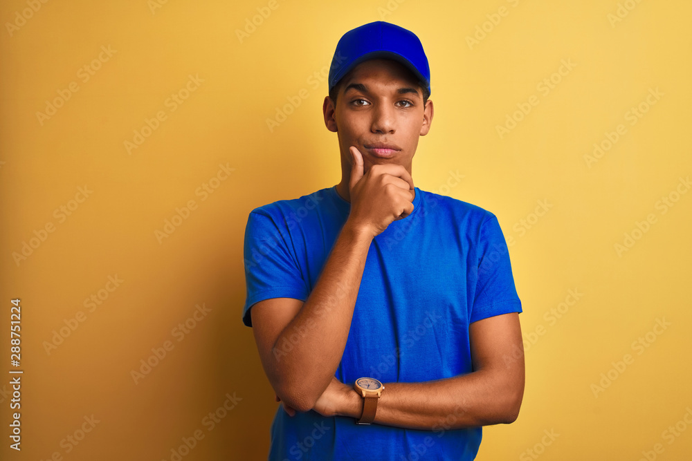 Young handsome arab delivery man standing over isolated yellow background looking confident at the camera with smile with crossed arms and hand raised on chin. Thinking positive.