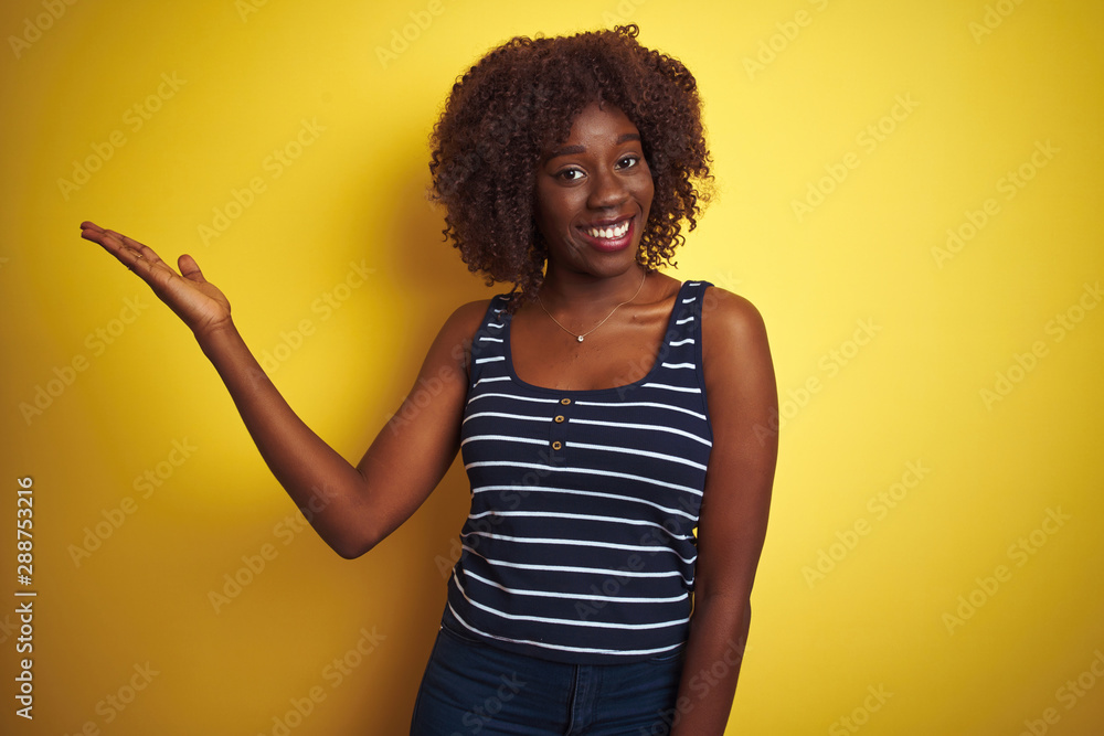 Young african afro woman wearing striped t-shirt over isolated yellow background smiling cheerful presenting and pointing with palm of hand looking at the camera. Young african afro woman wearing striped t-shirt over isolated yellow background smiling cheerful presenting and pointing with palm of hand looking at the camera.