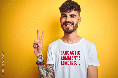 Young man with tattoo wearing fanny t-shirt standing over isolated yellow background smiling with happy face winking at the camera doing victory sign. Number two.