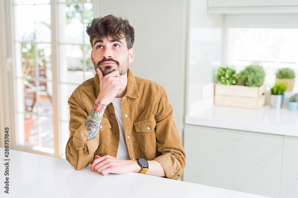 Young man wearing casual jacket sitting on white table with hand on chin thinking about question, pensive expression. Smiling with thoughtful face. Doubt concept.