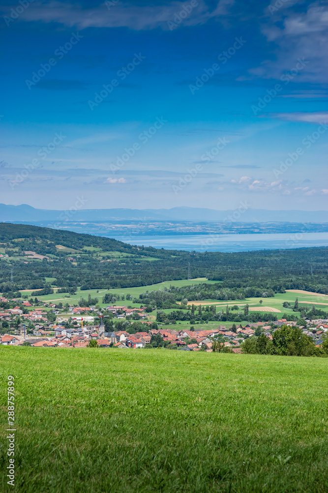  City landscape in the mountains, hills, fields, forests, green meadows, lakes in the distance and blue sky with clouds,focus area in the city.Town Bons-en-Chablais, Haute-Savoie in France.