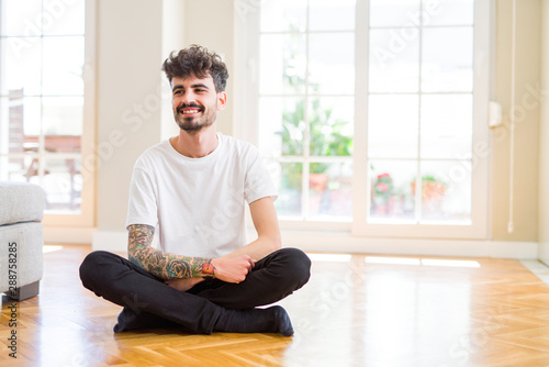 Young man working sitting casual on the floor at home looking away to side with smile on face, natural expression. Laughing confident.