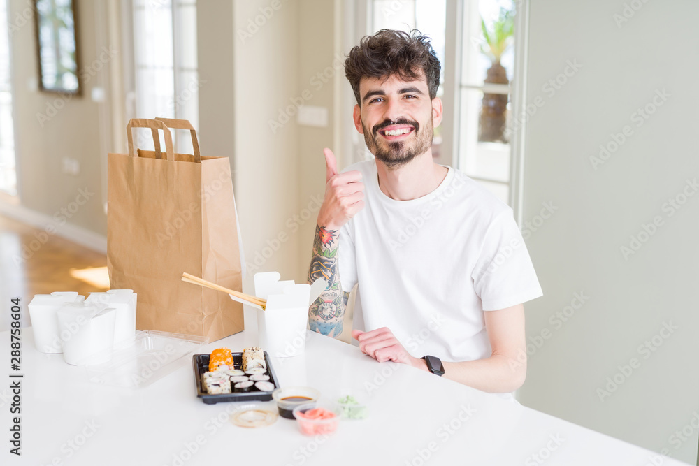 Young man eating asian sushi from home delivery doing happy thumbs up gesture with hand. Approving expression looking at the camera with showing success. Young man eating asian sushi from home delivery doing happy thumbs up gesture with hand. Approving expression looking at the camera with showing success.