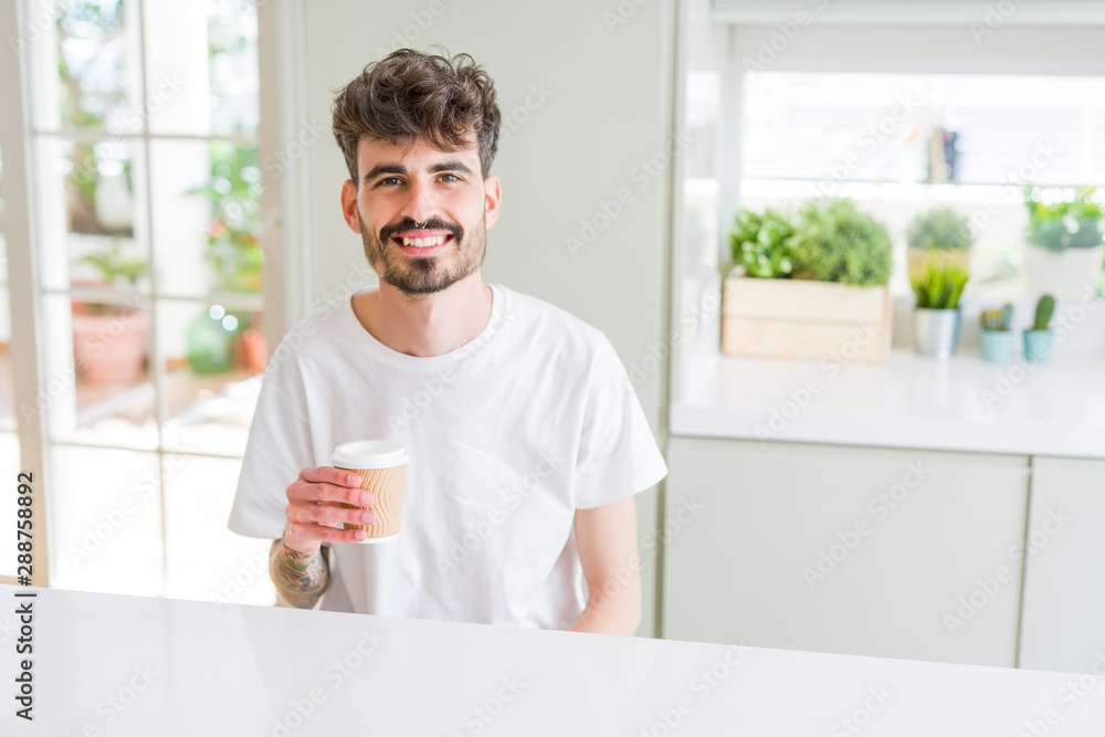 Young man drinking a coffee in a paper cup in the morning with a happy face standing and smiling with a confident smile showing teeth