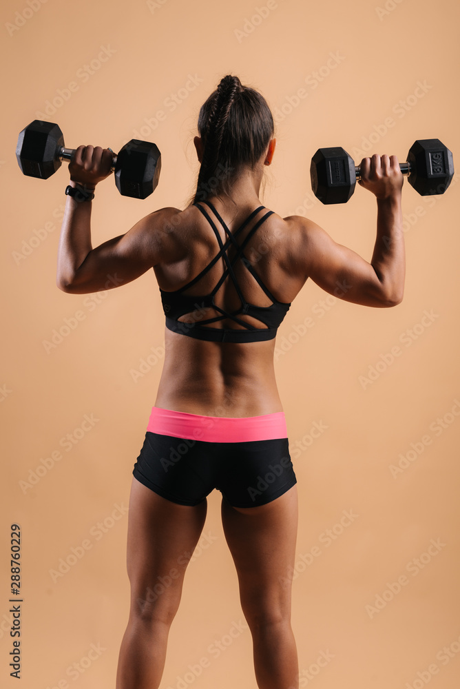 Back view portrait of beautiful fitness female with perfect muscular body in sports black bra. Sporty muscular girl is lifting dumbbells at shoulder level on isolated light red background
