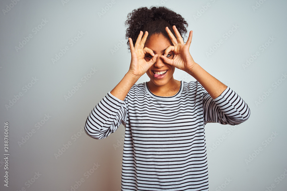African american woman wearing navy striped t-shirt standing over isolated white background doing ok gesture like binoculars sticking tongue out, eyes looking through fingers. Crazy expression. African american woman wearing navy striped t-shirt standing over isolated white background doing ok gesture like binoculars sticking tongue out, eyes looking through fingers. Crazy expression.