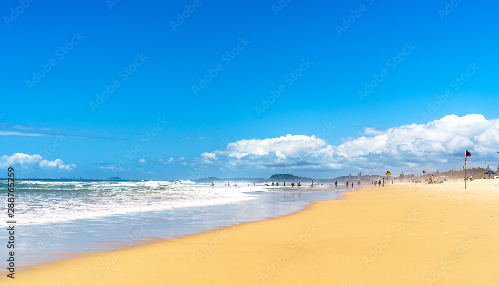 Beautiful wide panoramic view of the ocean waves rolling towards tropical sandy beach and silhouettes of surfers and beachgoers relaxing in the distance.