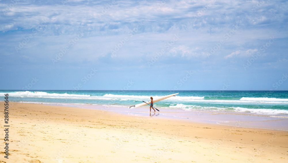 A young man walking along the ocean with his paddle board, with beautiful blue sky and waves on the background.