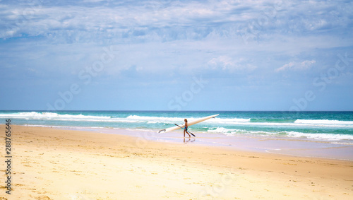 A young man walking along the ocean with his paddle board, with beautiful blue sky and waves on the background.