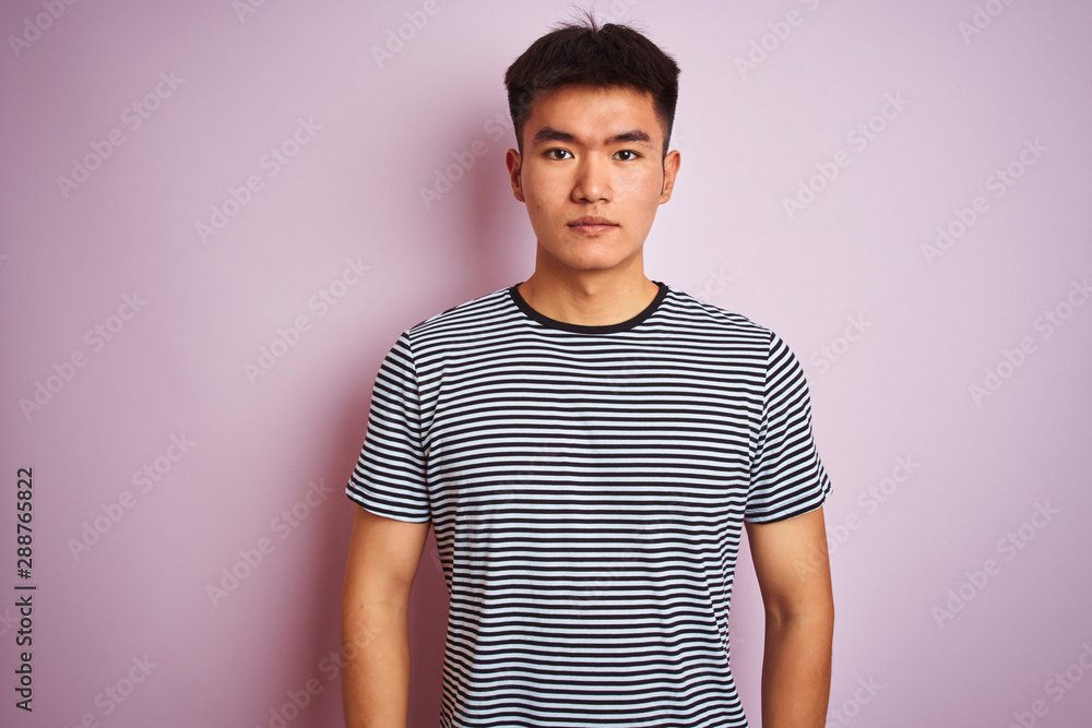 Young asian chinese man wearing striped t-shirt standing over isolated pink background with serious expression on face. Simple and natural looking at the camera. Young asian chinese man wearing striped t-shirt standing over isolated pink background with serious expression on face. Simple and natural looking at the camera.