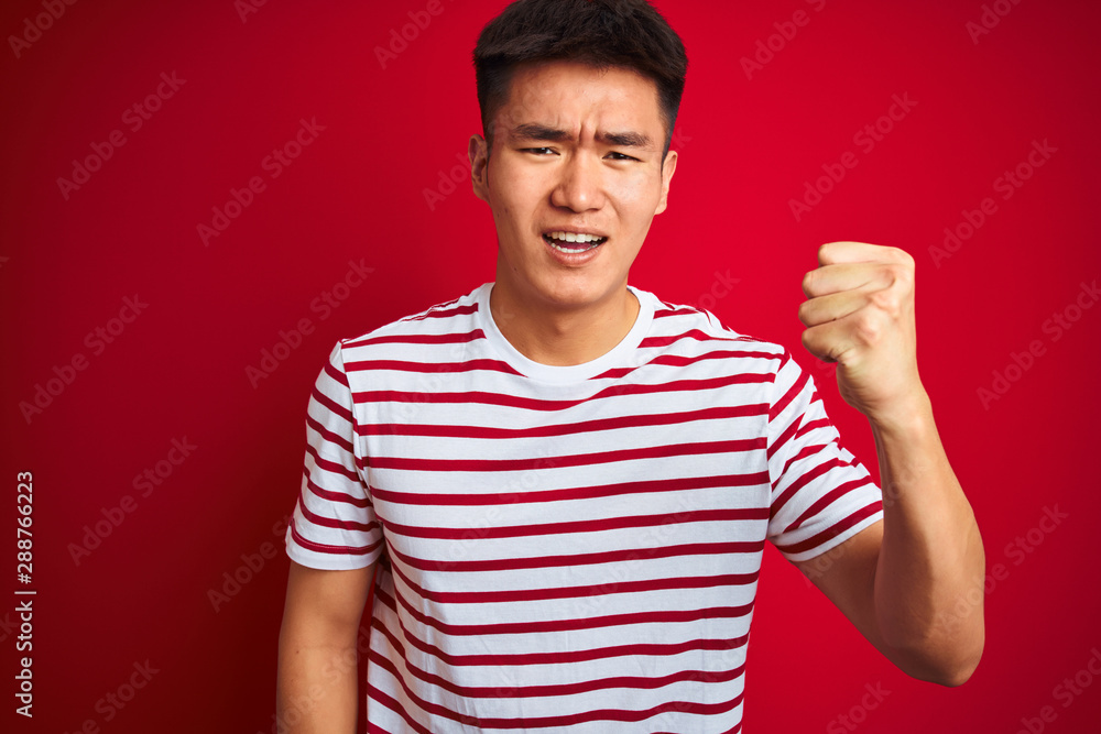 Young asian chinese man wearing striped t-shirt standing over isolated red background angry and mad raising fist frustrated and furious while shouting with anger. Rage and aggressive concept. Young asian chinese man wearing striped t-shirt standing over isolated red background angry and mad raising fist frustrated and furious while shouting with anger. Rage and aggressive concept.