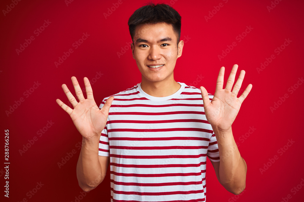 Young asian chinese man wearing striped t-shirt standing over isolated red background showing and pointing up with fingers number ten while smiling confident and happy. Young asian chinese man wearing striped t-shirt standing over isolated red background showing and pointing up with fingers number ten while smiling confident and happy.