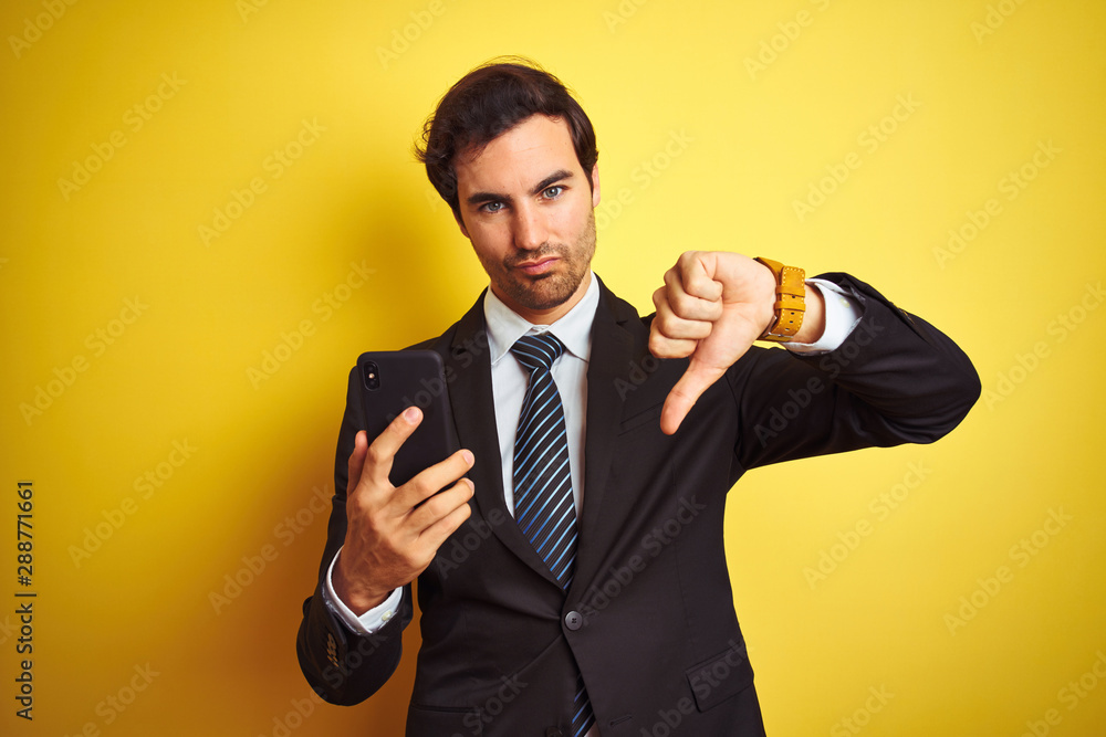 Young handsome businessman using smartphone standing over isolated yellow background with angry face, negative sign showing dislike with thumbs down, rejection concept Young handsome businessman using smartphone standing over isolated yellow background with angry face, negative sign showing dislike with thumbs down, rejection concept