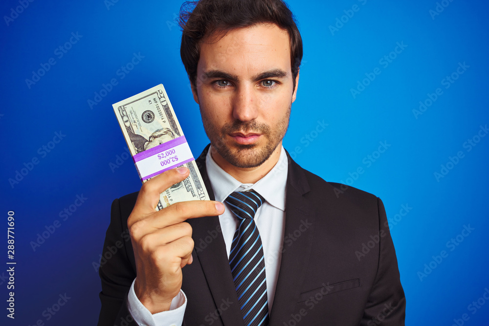 Young handsome businessman wearing suit holding dollars over isolated blue background with a confident expression on smart face thinking serious Young handsome businessman wearing suit holding dollars over isolated blue background with a confident expression on smart face thinking serious