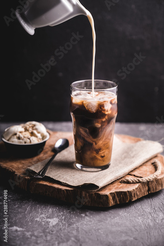  Iced coffee with milk. Preparation of iced coffee. Shot with streaks of milk in black coffee on dark concrete background and wooden tray.