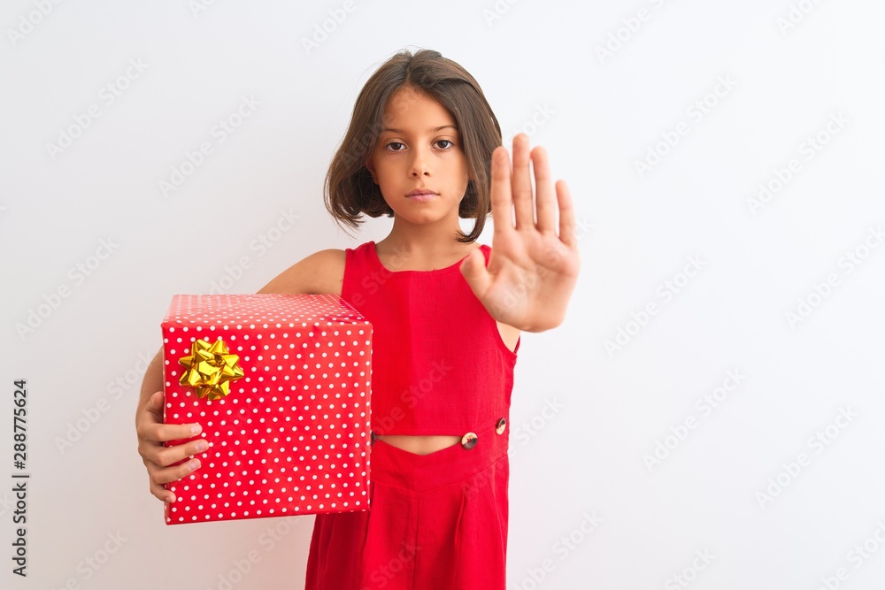 Beautiful child girl holding birthday gift standing over isolated white background with open hand doing stop sign with serious and confident expression, defense gesture Beautiful child girl holding birthday gift standing over isolated white background with open hand doing stop sign with serious and confident expression, defense gesture