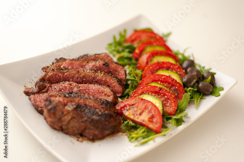 Cut-up grilled beef steak on white plate on white table with salad