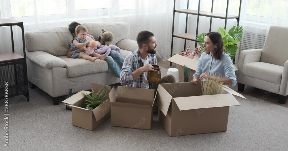 Parents unpacking carton box while kids are playing on sofa in new house Parents unpacking carton box while kids are playing on sofa in new house