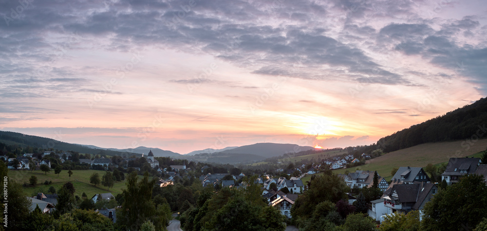 Wide panoramic view over the mountainous village of Grafschaft in the winter sports region of Sauerland, Germany, during sunset with a partly colourful orange lit cloudy sky over the valley
