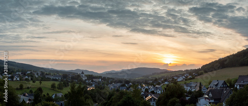 Wide panoramic view over the mountainous village of Grafschaft in the winter sports region of Sauerland, Germany, during sunset with a partly colourful orange lit cloudy sky © Maarten Zeehandelaar