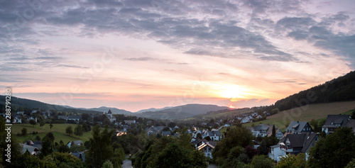 Wide panoramic view over the mountainous village of Grafschaft in the winter sports region of Sauerland, Germany, during sunset with a partly colourful orange lit cloudy sky over the valley