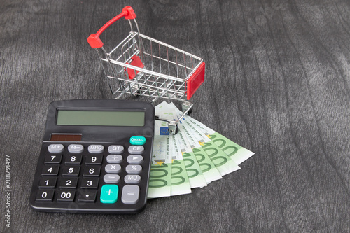 Shopping Cart and calculator. Purchase Cost Calculation. Shopping consept, euro money, calculator and cart. Shopping cart with euro on grey desk. Copy space for text.