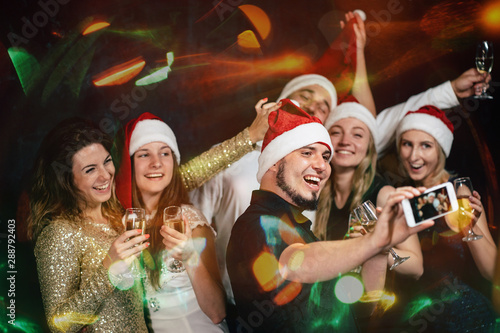 New Year, event, party, holiday, fun. Company of cheerful friends in Santa caps celebrating, making selfie on smart phone, drinking champagne