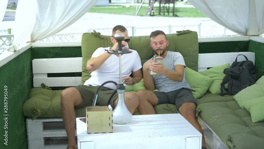 two friends smoke a hookah and watch the application on the phone while sitting in the gazebo outdoors on a sunny summer day.