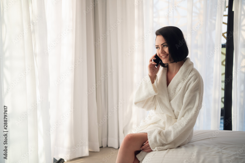Woman wearing white bathrobe in the morning near window in the light room. Talking by the phone Woman wearing white bathrobe in the morning near window in the light room. Talking by the phone