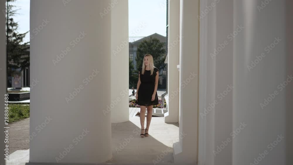 Gorgeous beautiful blonde in a black dress on a walk passes among the white columns of the building. Steadicam shot. Gorgeous beautiful blonde in a black dress on a walk passes among the white columns of the building. Steadicam shot.