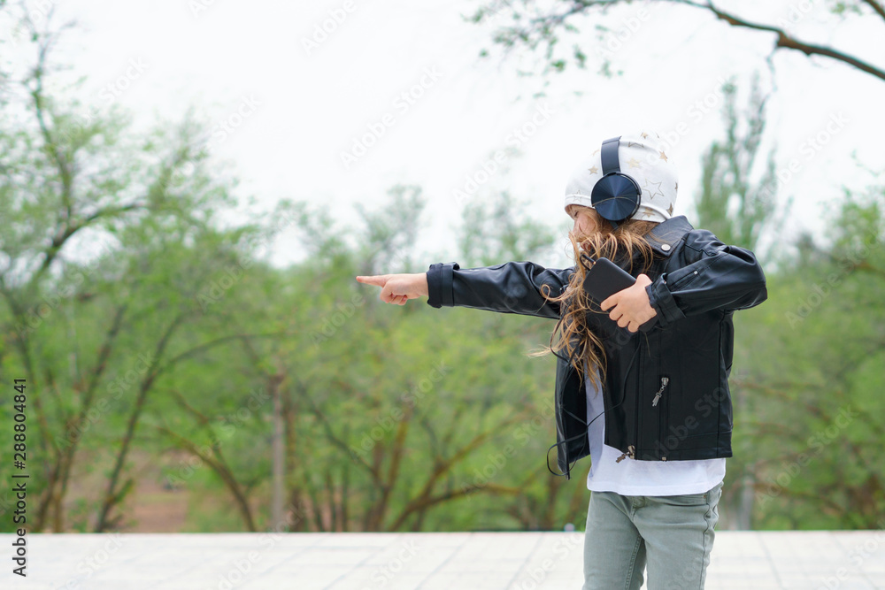 Girl has fun on street. She points direction, wears white T-shirt and black leather jacket. Schoolchild listening to energy music with headphones. Expressing true positive emotions of entertainment